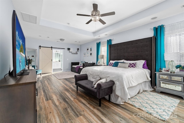 bedroom with a barn door, hardwood / wood-style flooring, ceiling fan, a tray ceiling, and multiple windows