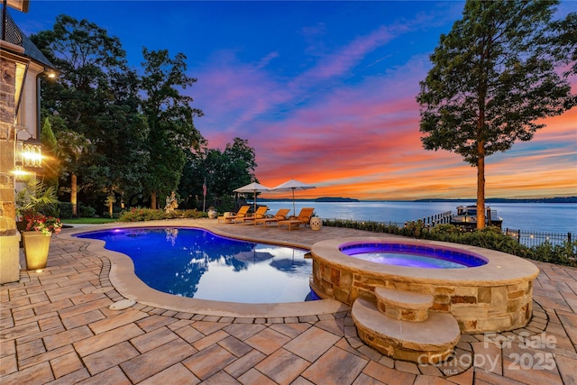 pool at dusk with an in ground hot tub, a water view, and a patio