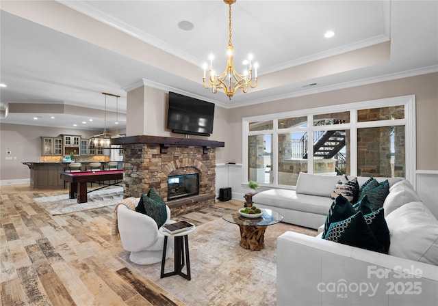 living room with a stone fireplace, crown molding, light wood-type flooring, a tray ceiling, and a chandelier