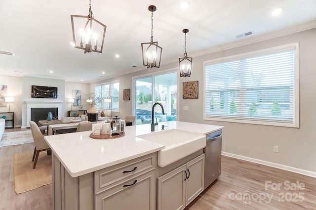 kitchen featuring an island with sink, sink, hanging light fixtures, stainless steel dishwasher, and light wood-type flooring