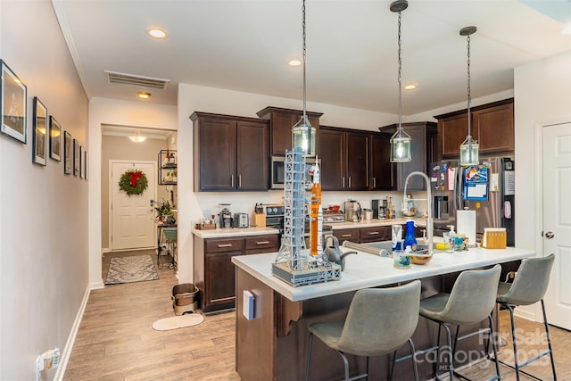 kitchen with appliances with stainless steel finishes, dark brown cabinets, pendant lighting, a center island with sink, and light hardwood / wood-style floors