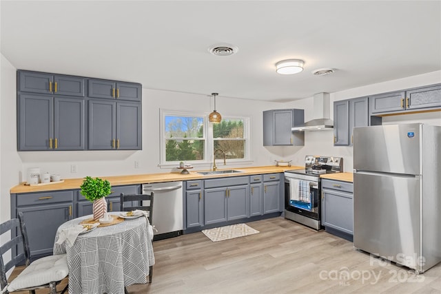 kitchen featuring wood counters, appliances with stainless steel finishes, wall chimney range hood, sink, and hanging light fixtures