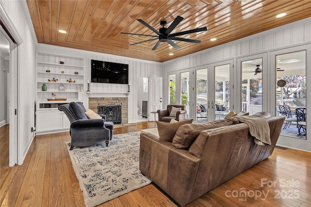 living room with built in shelves, ceiling fan, french doors, a stone fireplace, and wood ceiling