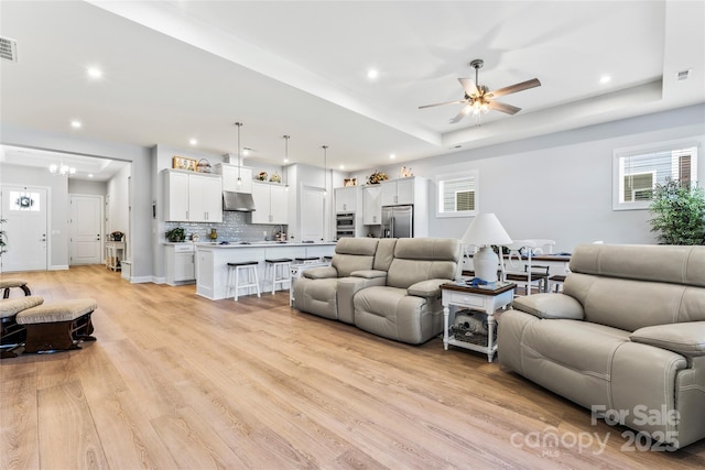 living room with ceiling fan with notable chandelier, a raised ceiling, and light hardwood / wood-style floors