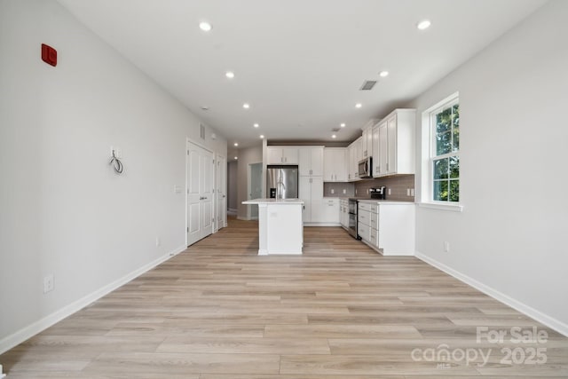 kitchen featuring light wood-type flooring, backsplash, stainless steel appliances, a center island with sink, and white cabinets