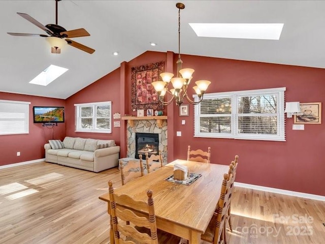 dining room featuring a fireplace, ceiling fan with notable chandelier, light hardwood / wood-style floors, and vaulted ceiling with skylight