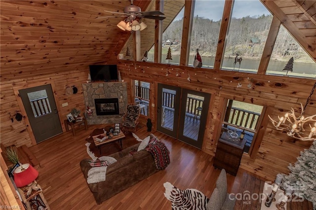 living room featuring high vaulted ceiling, ceiling fan with notable chandelier, wooden walls, a fireplace, and wood-type flooring