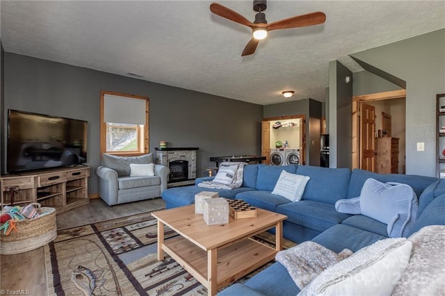 living room with ceiling fan, a stone fireplace, washing machine and dryer, a textured ceiling, and light wood-type flooring