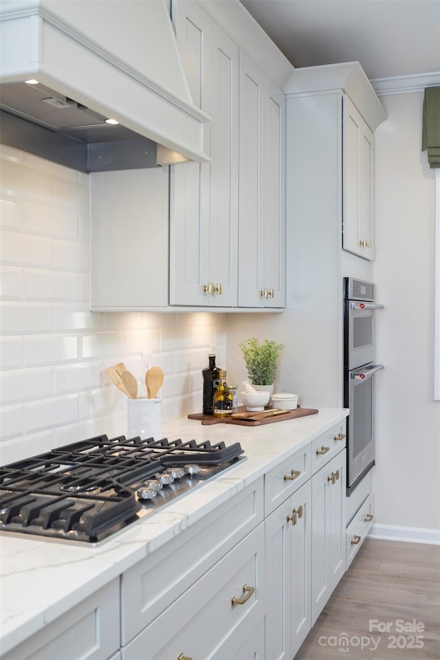 kitchen with custom range hood, stainless steel appliances, light wood-style floors, white cabinetry, and backsplash