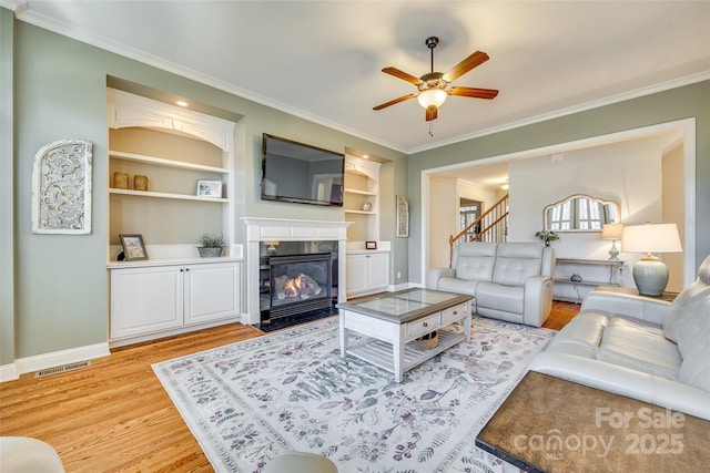 living room featuring light wood-type flooring, built in shelves, ceiling fan, crown molding, and a tiled fireplace