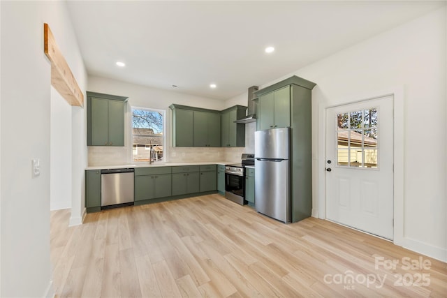 kitchen featuring stainless steel appliances, wall chimney range hood, green cabinetry, decorative backsplash, and light wood-type flooring