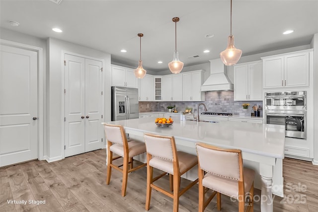 kitchen with a center island with sink, appliances with stainless steel finishes, white cabinetry, and custom range hood