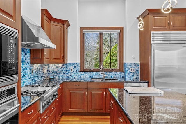 kitchen with backsplash, dark stone counters, wall chimney range hood, sink, and built in appliances