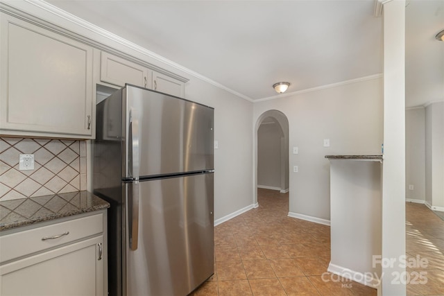 kitchen featuring dark stone countertops, light tile patterned floors, decorative backsplash, stainless steel refrigerator, and crown molding