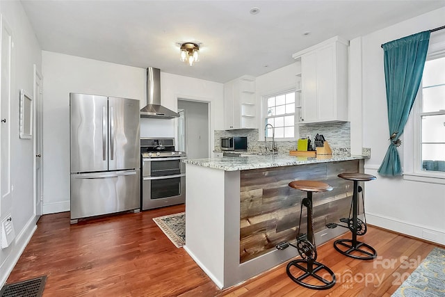 kitchen with kitchen peninsula, wall chimney exhaust hood, a kitchen bar, white cabinetry, and stainless steel appliances