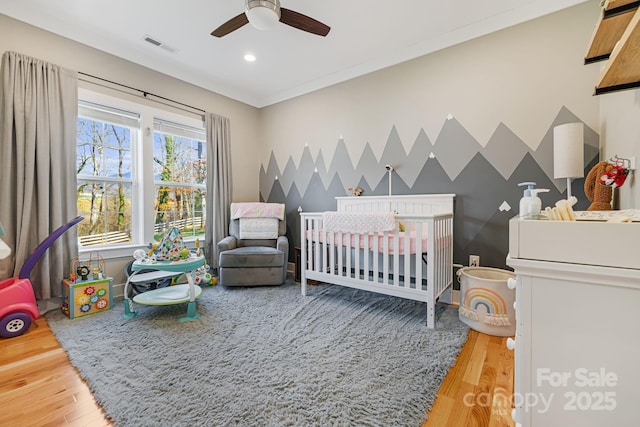 bedroom featuring wood-type flooring, a nursery area, ornamental molding, and ceiling fan