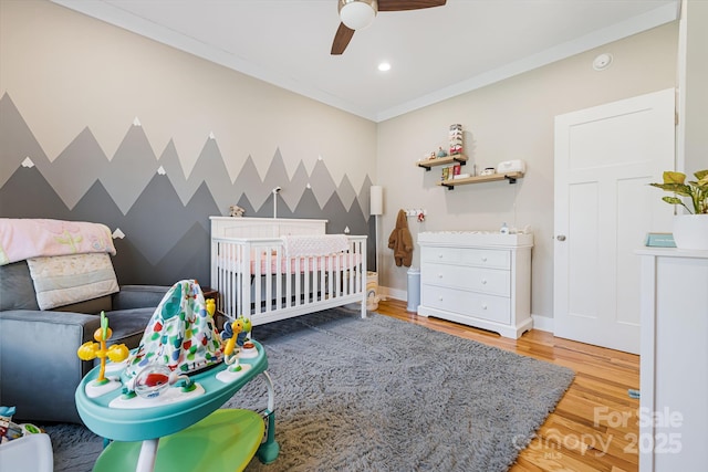 bedroom featuring ceiling fan, hardwood / wood-style floors, a nursery area, and ornamental molding