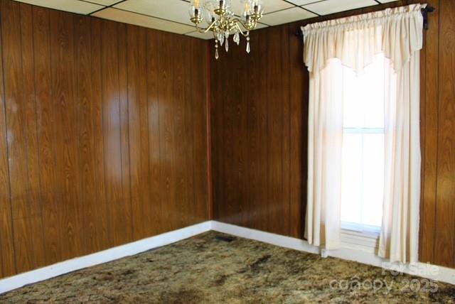 carpeted spare room featuring an inviting chandelier, a drop ceiling, and wooden walls