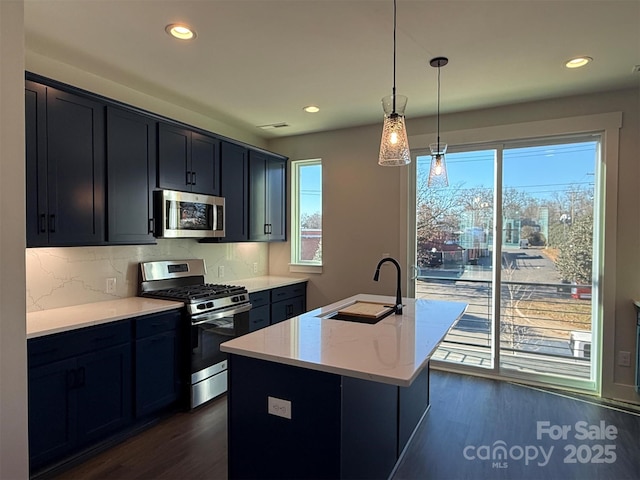 kitchen with light stone countertops, sink, hanging light fixtures, stainless steel appliances, and an island with sink