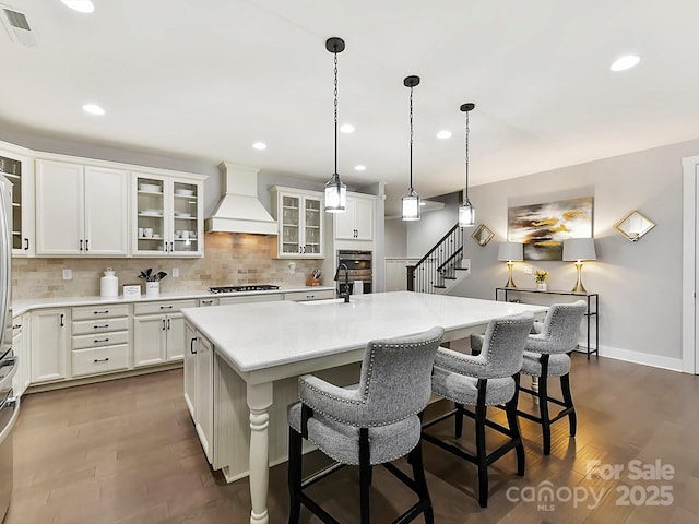 kitchen featuring custom exhaust hood, decorative backsplash, an island with sink, appliances with stainless steel finishes, and white cabinetry