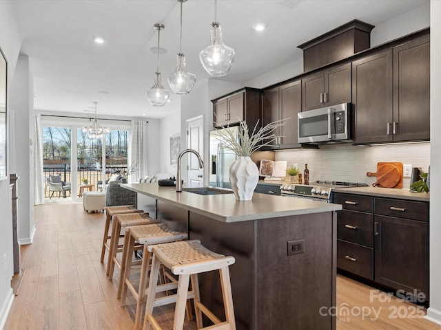 kitchen with dark brown cabinetry, light wood-type flooring, decorative backsplash, stainless steel microwave, and a center island with sink