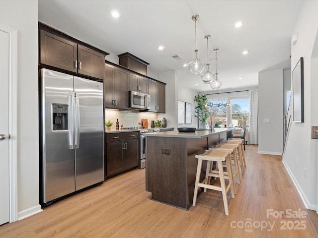 kitchen with dark brown cabinetry, decorative backsplash, an island with sink, appliances with stainless steel finishes, and light wood-style floors