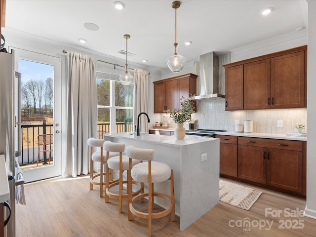 kitchen with a kitchen island with sink, wall chimney range hood, light wood-style floors, light countertops, and decorative backsplash