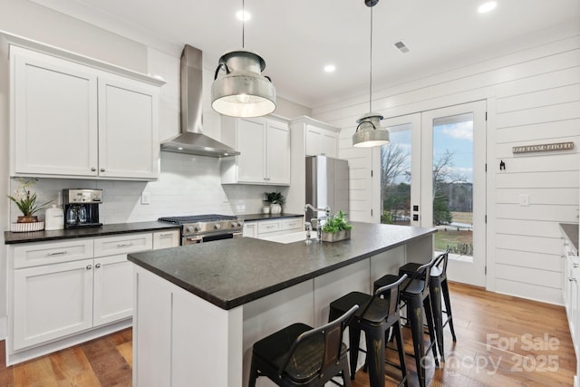 kitchen featuring decorative light fixtures, appliances with stainless steel finishes, an island with sink, white cabinets, and wall chimney exhaust hood