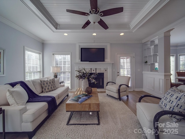 living room featuring a tray ceiling, a tiled fireplace, crown molding, and ceiling fan