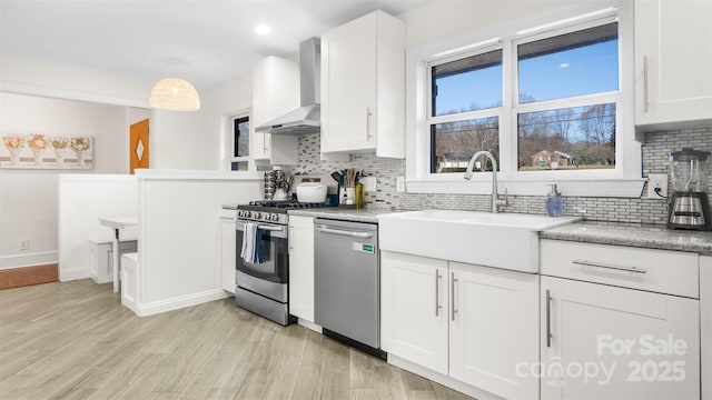 kitchen with appliances with stainless steel finishes, decorative light fixtures, white cabinetry, and wall chimney range hood