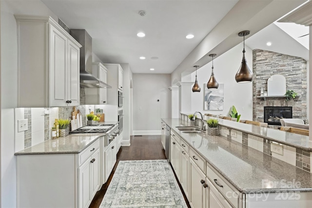 kitchen featuring wall chimney range hood, sink, hanging light fixtures, light stone counters, and white cabinetry