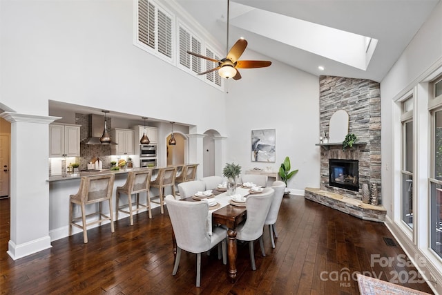 dining space featuring a skylight, ceiling fan, a stone fireplace, high vaulted ceiling, and dark hardwood / wood-style floors