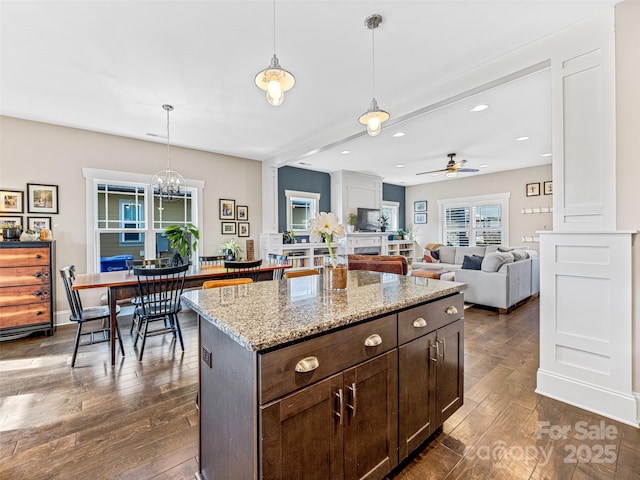 kitchen with light stone countertops, ceiling fan with notable chandelier, dark brown cabinetry, dark wood-type flooring, and pendant lighting