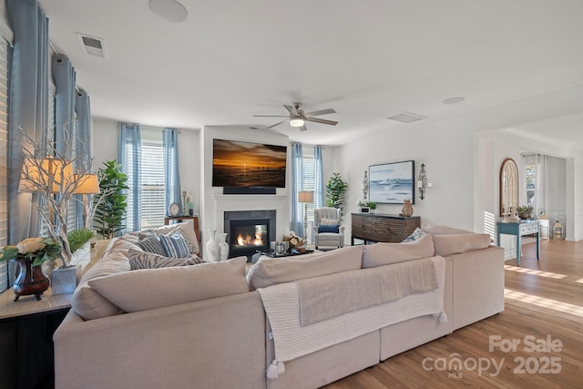 living room featuring light wood-type flooring, a glass covered fireplace, visible vents, and ceiling fan