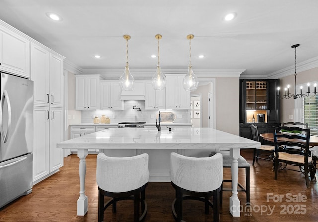 kitchen featuring stainless steel refrigerator, white cabinetry, hanging light fixtures, a kitchen island with sink, and light stone counters