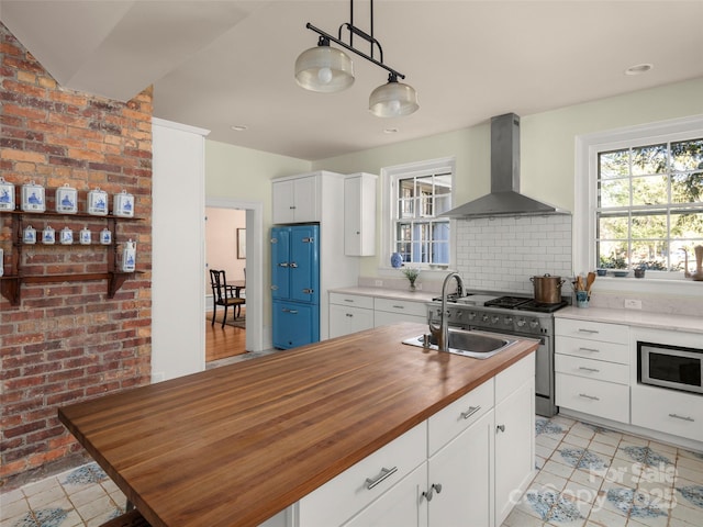 kitchen featuring hanging light fixtures, wall chimney range hood, wood counters, a kitchen island with sink, and white cabinets