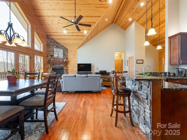 dining room with wooden ceiling, a stone fireplace, light hardwood / wood-style flooring, high vaulted ceiling, and ceiling fan with notable chandelier