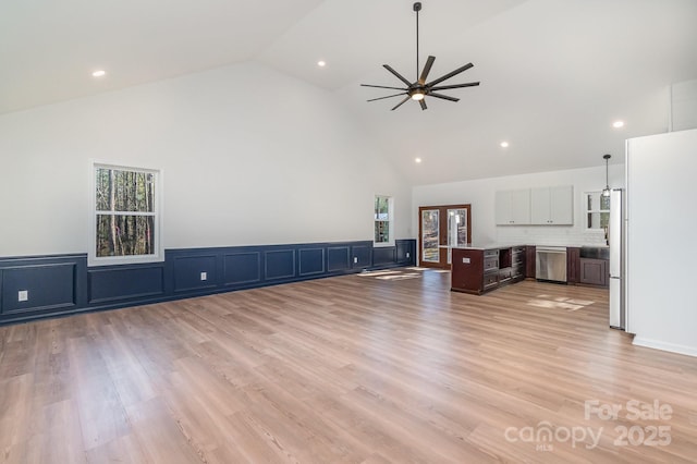 unfurnished living room featuring light wood-type flooring, high vaulted ceiling, a ceiling fan, and wainscoting