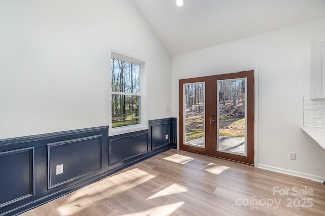 doorway to outside with lofted ceiling, a wainscoted wall, light wood-style flooring, french doors, and a decorative wall