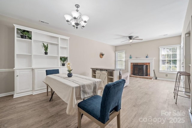 dining space featuring light wood-type flooring, visible vents, ceiling fan with notable chandelier, baseboards, and a brick fireplace