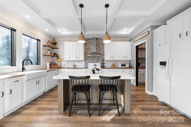 kitchen with white appliances, wall chimney range hood, a kitchen island, white cabinetry, and sink