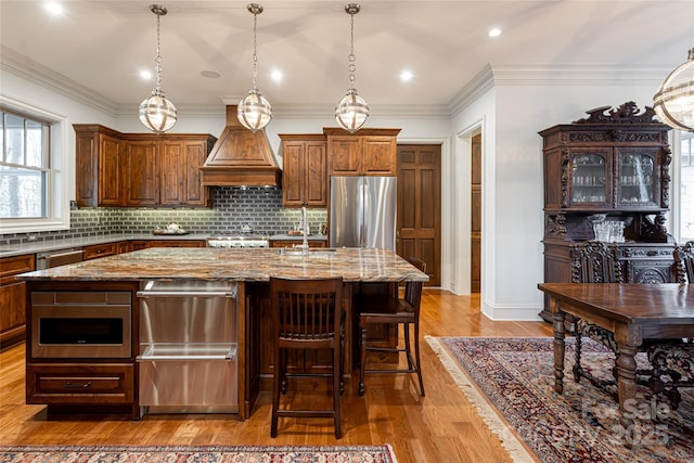 kitchen with light stone counters, tasteful backsplash, premium range hood, a kitchen island with sink, and appliances with stainless steel finishes