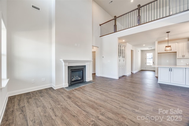 unfurnished living room featuring a fireplace with flush hearth, a towering ceiling, wood finished floors, visible vents, and baseboards