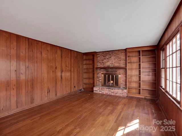 unfurnished living room featuring built in shelves, wooden walls, hardwood / wood-style floors, and a brick fireplace