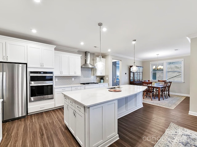 kitchen featuring a center island with sink, stainless steel appliances, hanging light fixtures, wall chimney exhaust hood, and white cabinetry