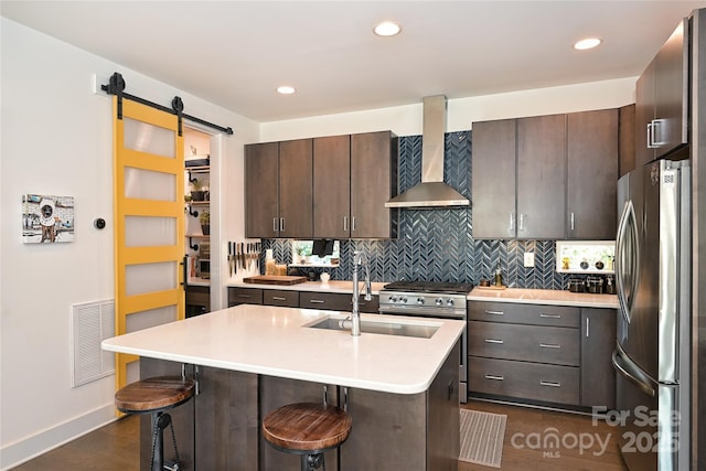 kitchen with stainless steel appliances, a barn door, a center island with sink, and wall chimney range hood