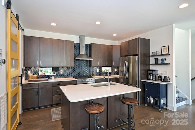 kitchen featuring wall chimney exhaust hood, sink, dark brown cabinets, appliances with stainless steel finishes, and an island with sink