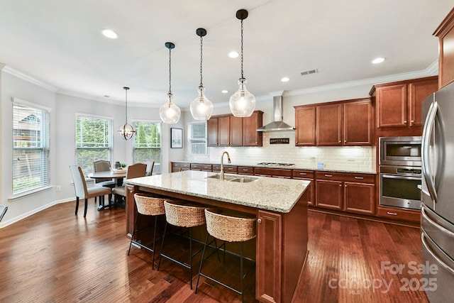 kitchen with sink, wall chimney range hood, pendant lighting, a center island with sink, and appliances with stainless steel finishes