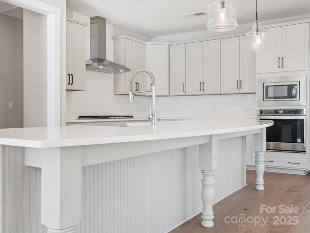 kitchen with wall chimney exhaust hood, stainless steel appliances, backsplash, a kitchen island with sink, and light wood-type flooring