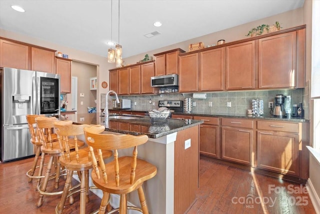 kitchen featuring stainless steel appliances, dark stone countertops, sink, hanging light fixtures, and a kitchen island with sink